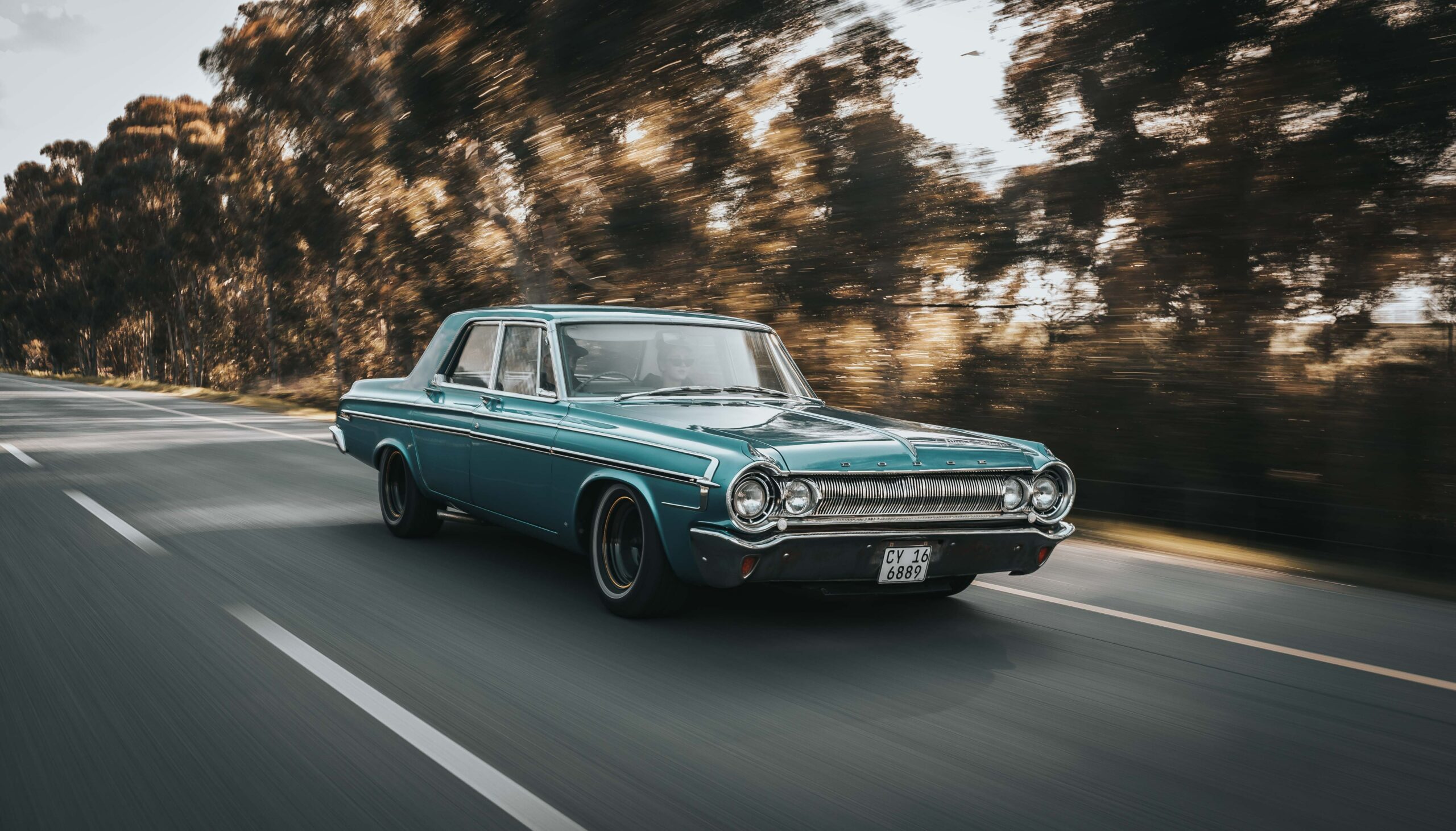 Teal colored muscle car on the road with trees in the background.