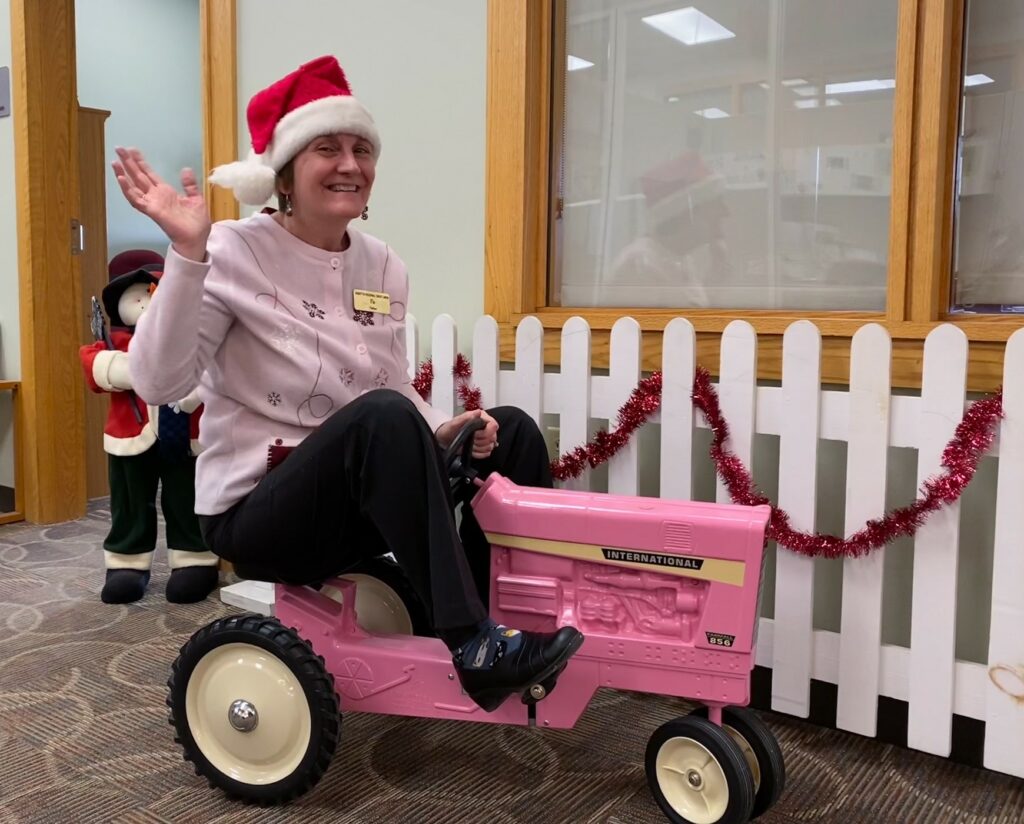 Woman wearing Santa hat and sitting on pink child's tractor.