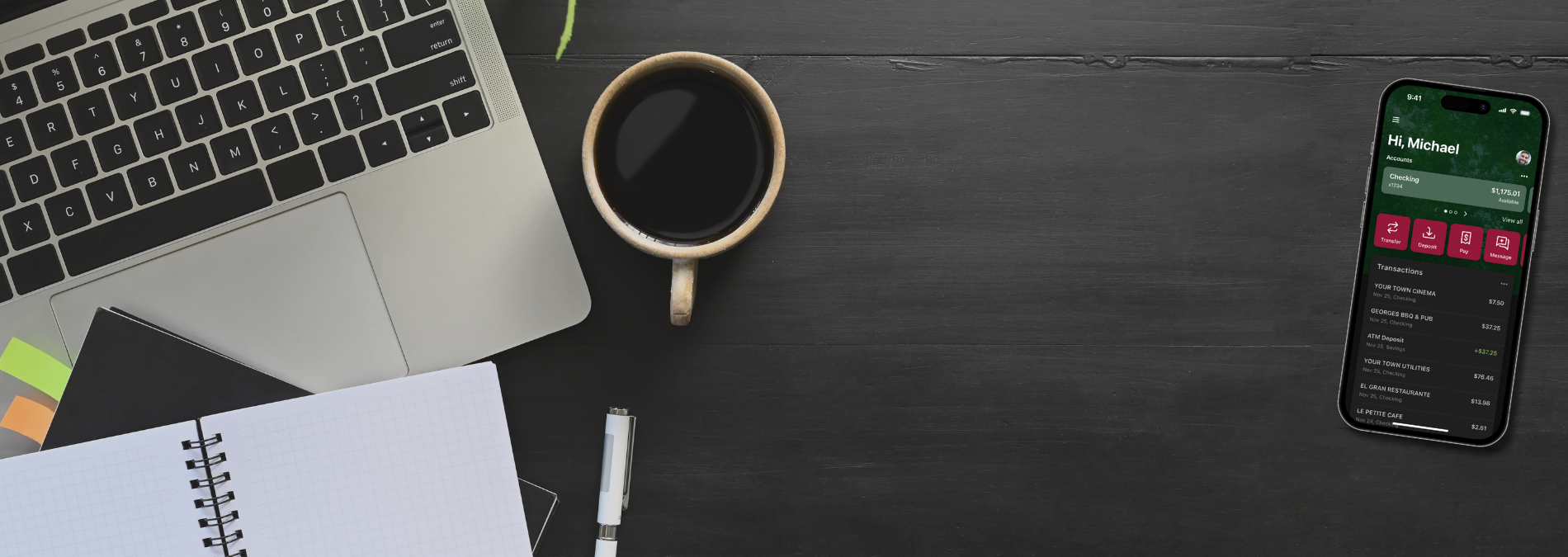 Desktop showing notebook, laptop computer, a mug of black coffee, holding phone displaying digital banking.