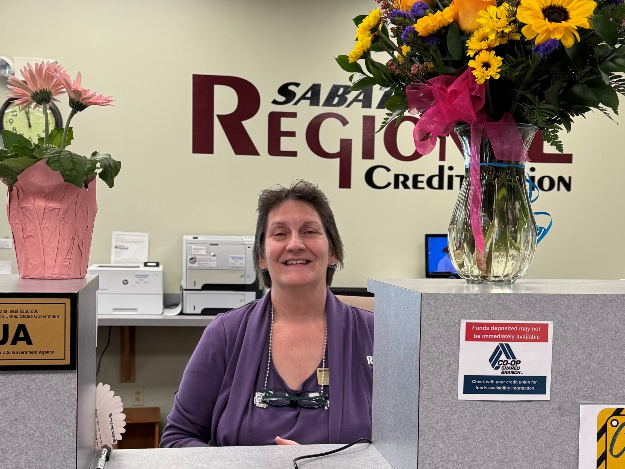 Mature woman bank teller standing at counter with a vase of flowers on each side of her.